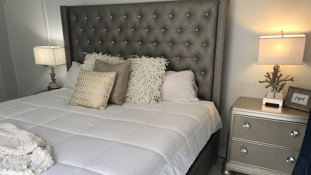 A neatly made bed with a tufted headboard, white bedding, and decorative pillows. There is a bedside table with a lamp and a small decorative item.