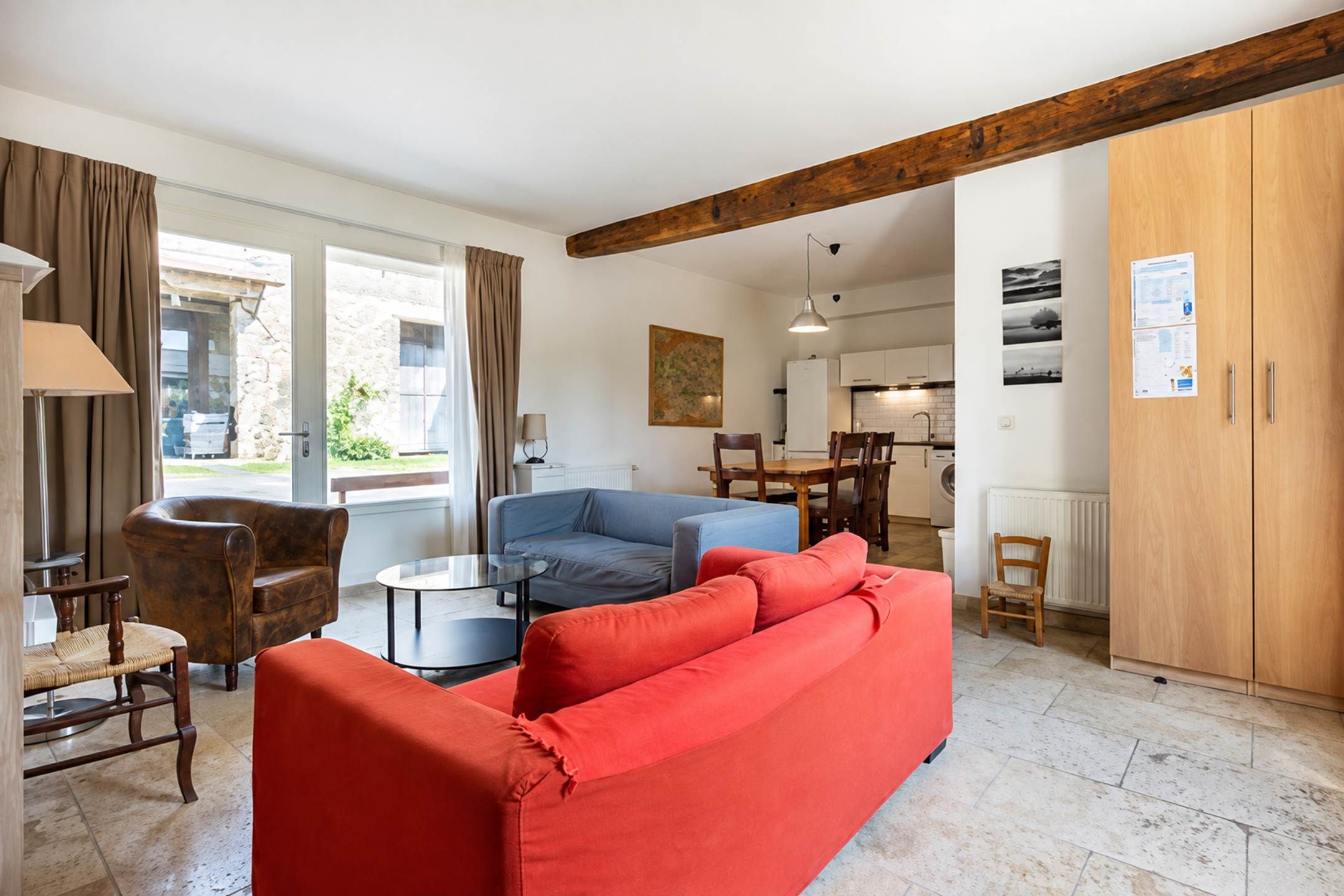 A living room with a red sofa, a blue armchair, and a small round table. There is a wooden beam ceiling and a view of the kitchen through an open door.