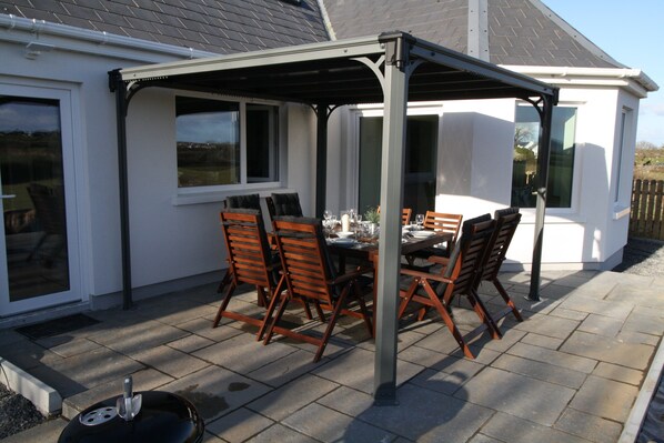 Een terras buiten met een tafel en stoelen, een overdekte zithoek en een wit gebouw met ramen.