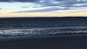 Una playa con vista al océano y a la costa distante bajo un cielo nublado.