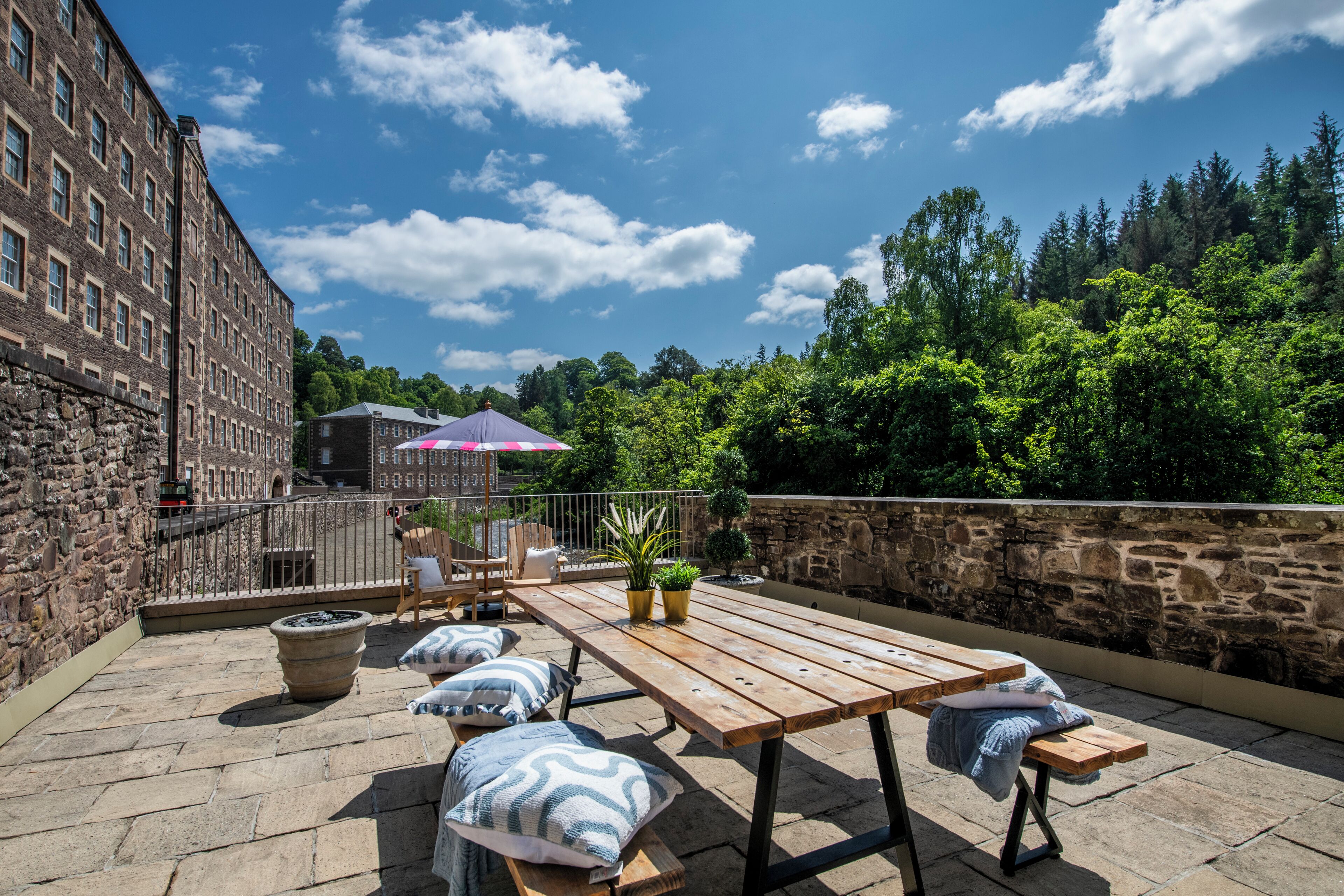 An outdoor patio with a wooden table and benches, surrounded by stone walls and a view of trees and a building.