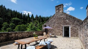 Un bâtiment en pierre doté d’une terrasse en bois, d’un espace extérieur aménagé pour s’asseoir et offrant une vue sur une forêt.