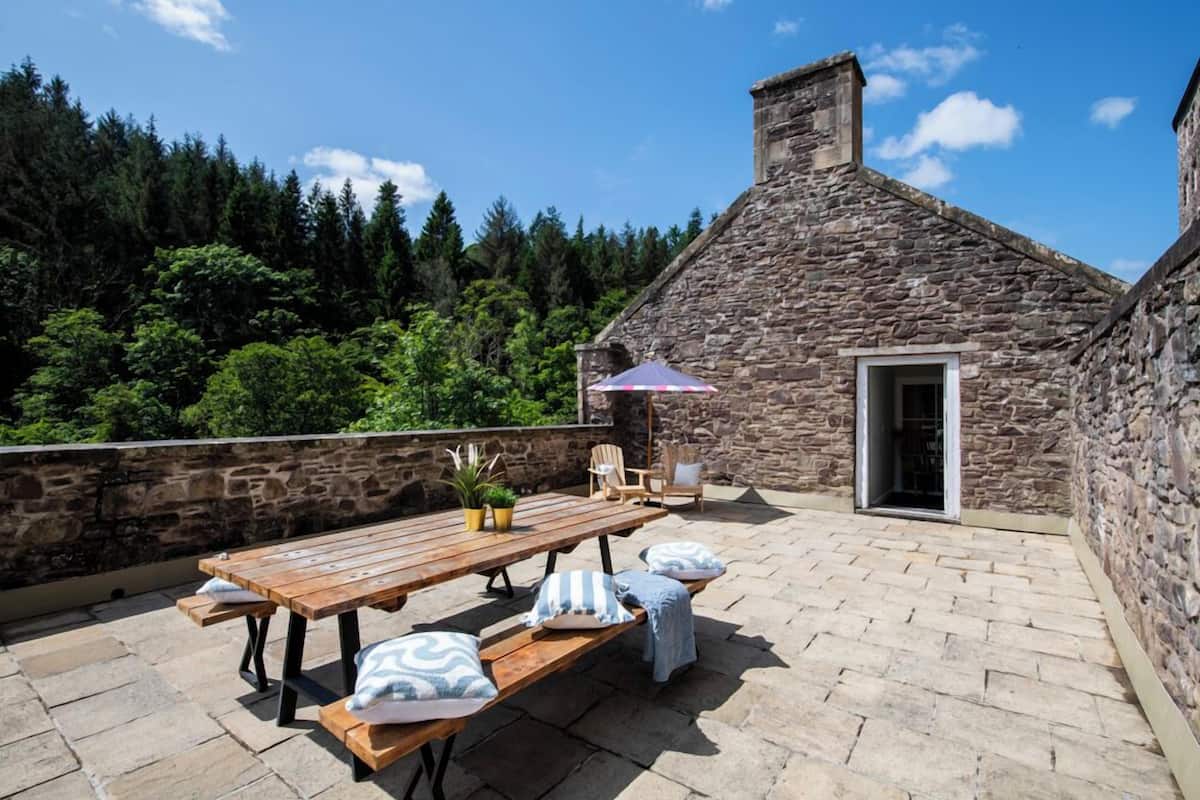 Un edificio de piedra con una terraza de madera, asientos al aire libre y vistas a un bosque.