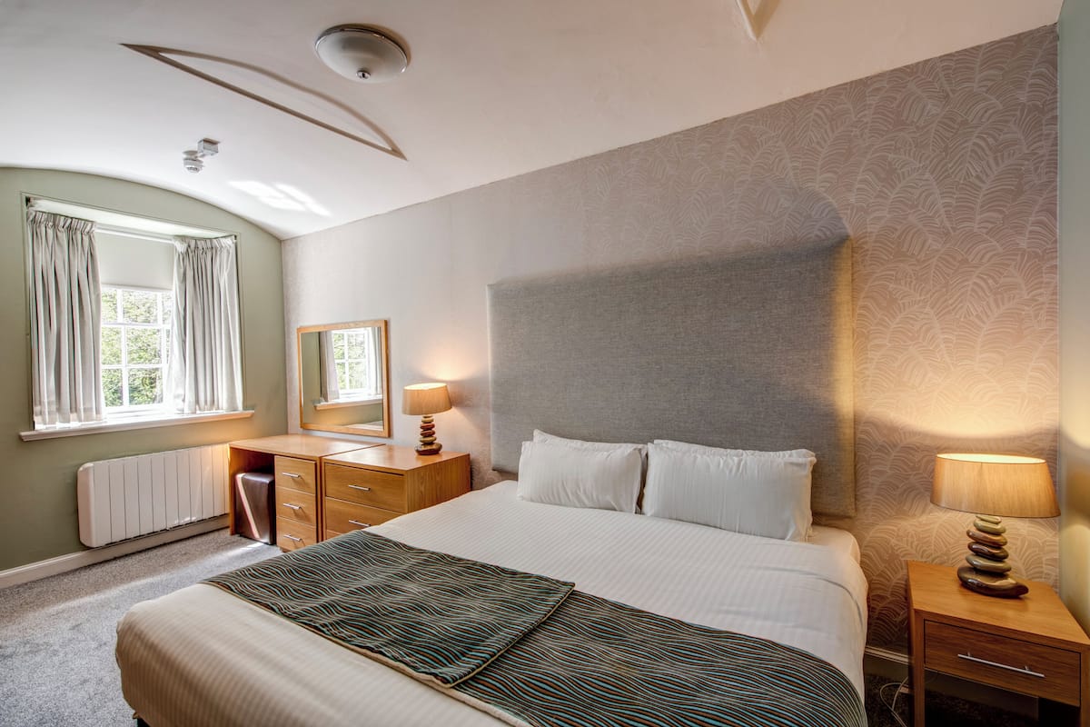 Una habitación de hotel con una cama grande, una mesita de noche con lámpara, un aparador, un espejo y una ventana con cortinas.