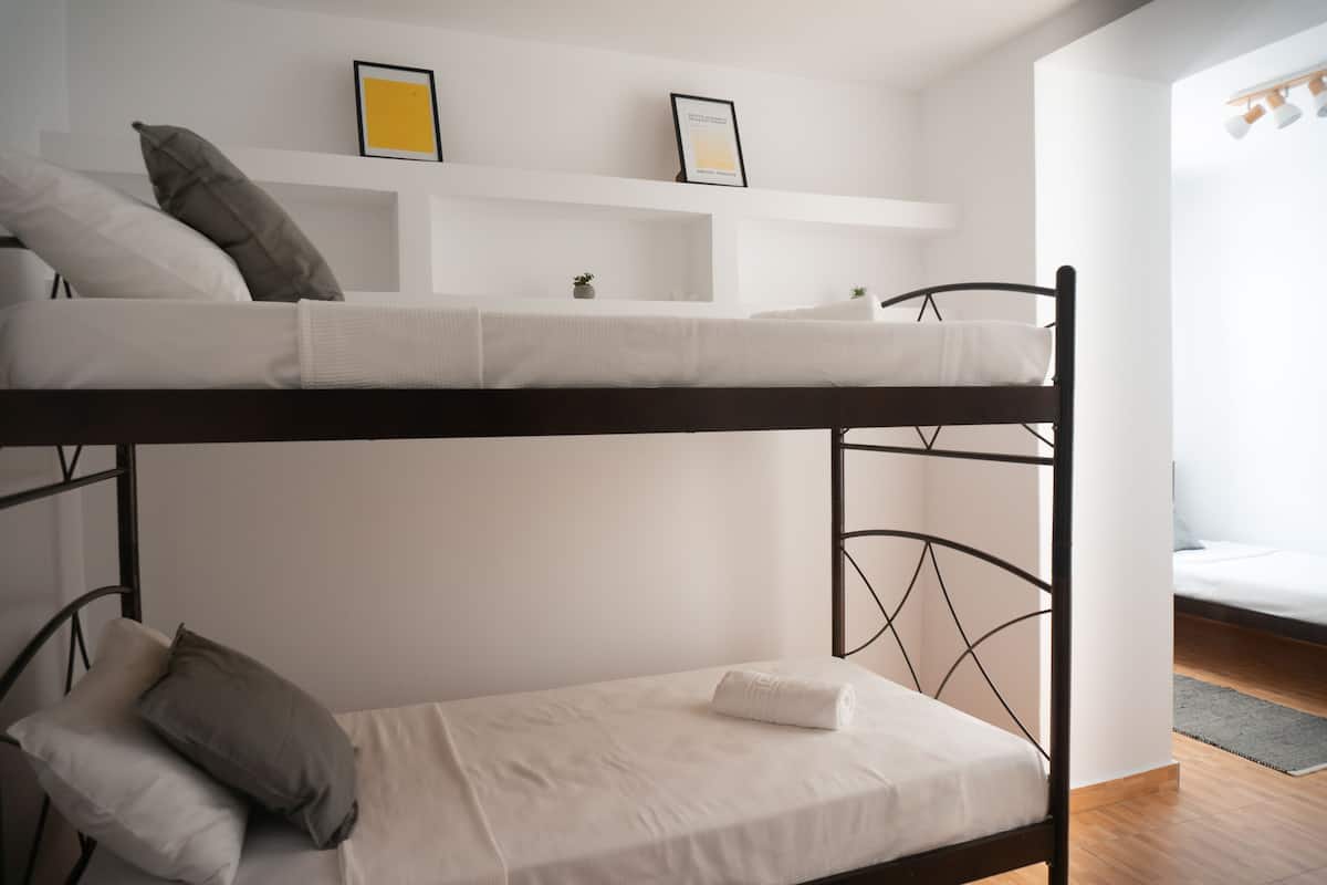 Dormitorio con literas, una pared blanca, un estante pequeño con dos cuadros enmarcados y una ventana con cortinas.