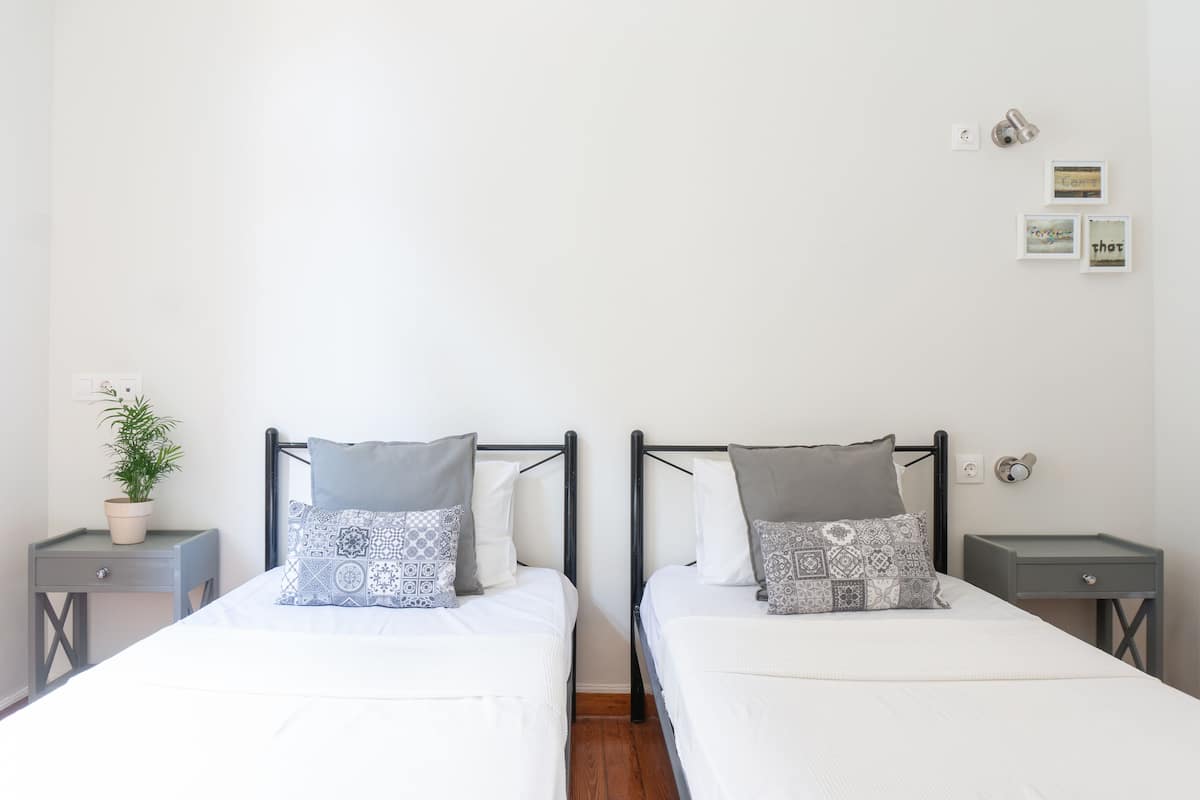 Habitación doble con dos camas, cada una con almohadas estampadas, una mesita de noche pequeña, una planta en maceta y un aplique de pared.