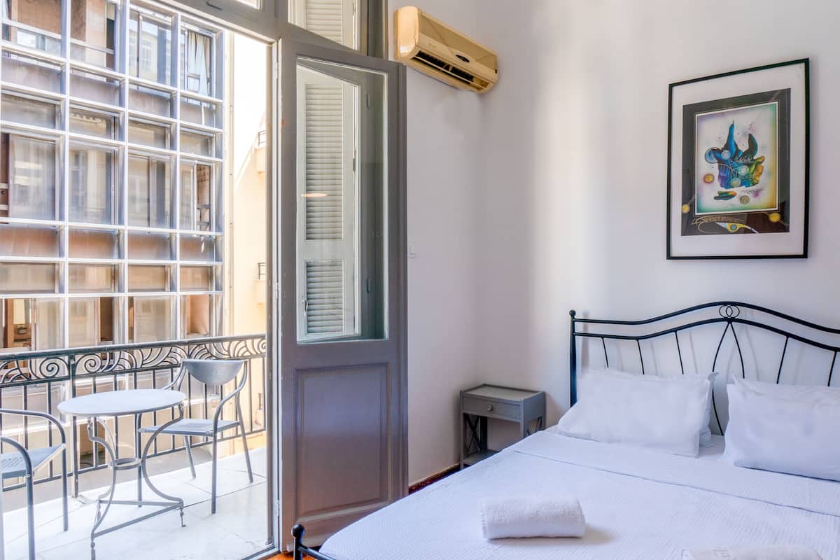 Una habitación de hotel con una cama, una mesita de noche pequeña, una silla, un balcón con mesa y sillas, y una ventana con persianas.
