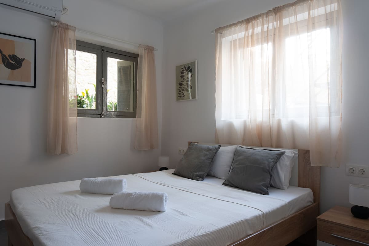 Una cama bien hecha con almohadas y una toalla enrollada, una mesita de noche con lámpara, y una ventana con cortinas traslúcidas.