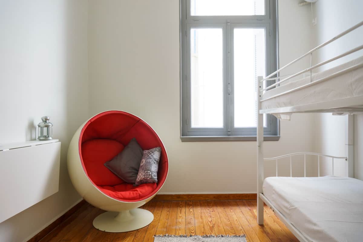 Una habitación con una cama litera, una silla roja con cojines, una ventana y un radiador.