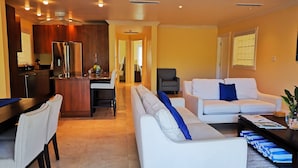 A modern living room with a white sofa, a dining area with a kitchen in the background, and a hallway leading to another room. - Sea Dreamer Villa 10E Hamilton Beach Villas and Spa -2BRD (Charlestown)
