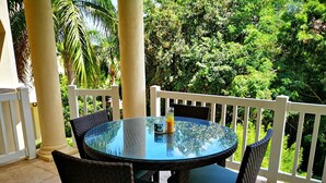 A round glass table with wicker chairs, a glass pitcher, and a glass of orange juice on a balcony with white railings and lush greenery in the background. - Sea Dreamer Villa 10E Hamilton Beach Villas and Spa -2BRD (Charlestown)