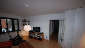 Ett modernt vardagsrum med trägolv, en röd soffa, ett träbord, en tv på ett tv-bord, en bokhylla och ett stort fönster.