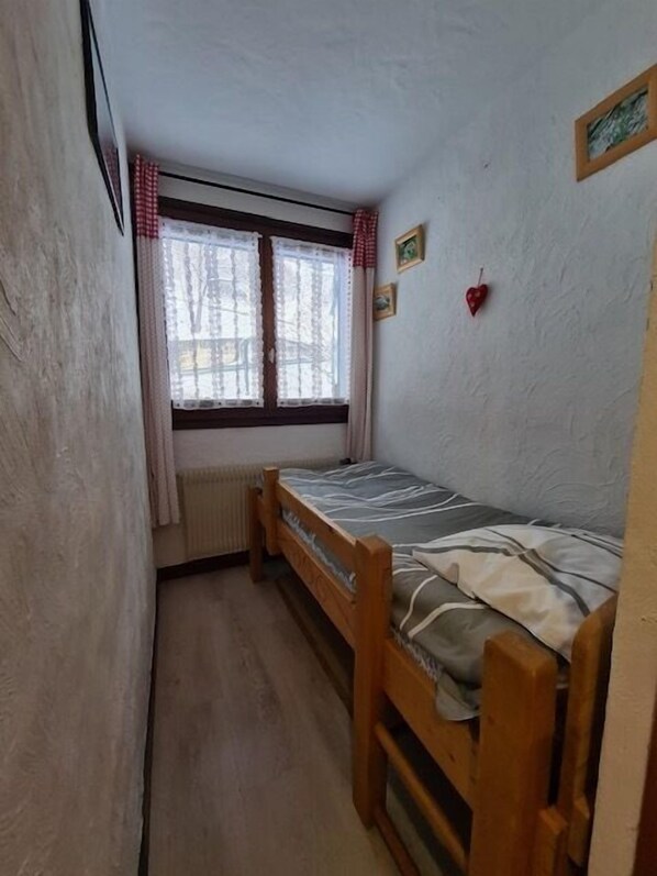A small, single-bed room with a wooden bed, a window with curtains, and a radiator. - Alpenrose (Saint-Gervais-les-Bains)
