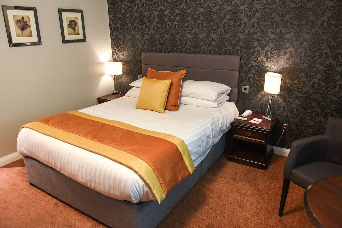 A neatly made bed with a brown and white bedspread, two bedside lamps, a nightstand with a phone and papers, and a chair.