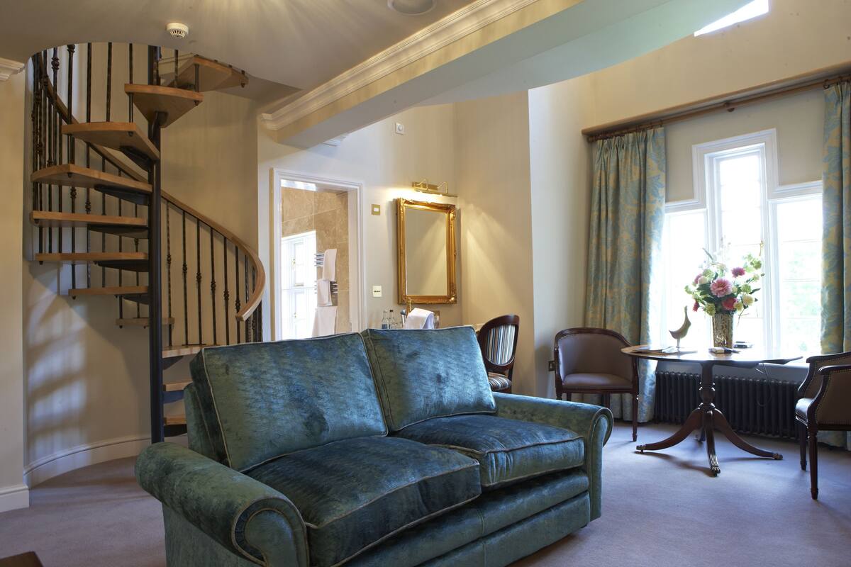 A living room with a green sofa, a wooden spiral staircase, a dining table with chairs, and a large mirror.