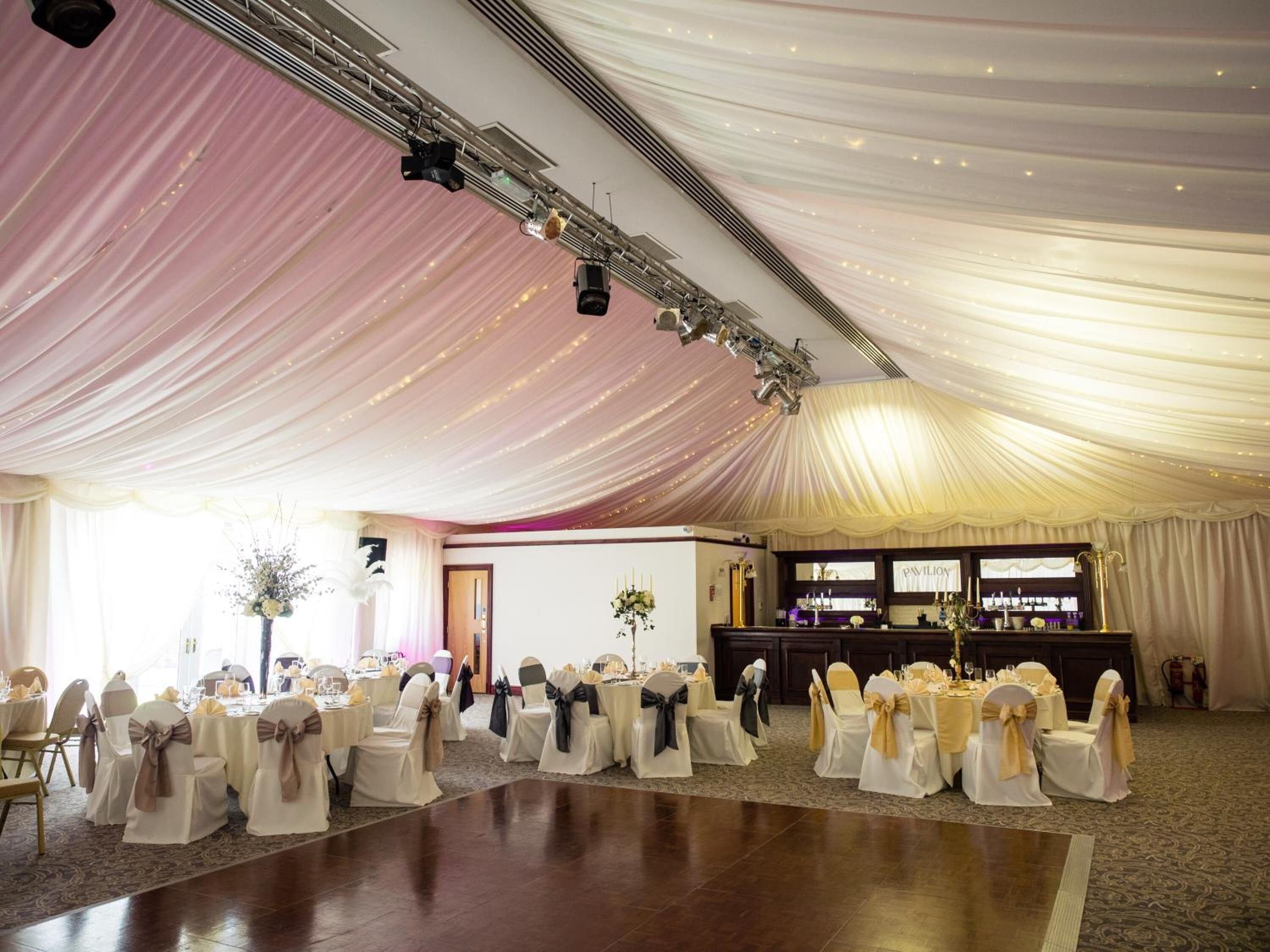 A banquet hall with tables set for a formal event, draped with white fabric and gold sashes.