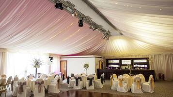A banquet hall with tables set for a formal event, draped with white fabric and gold sashes.