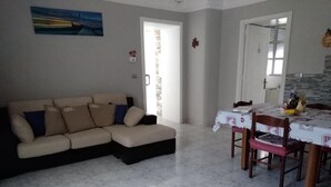 Una sala de estar con un sofá seccional color beige, una mesa de comedor con sillas y piso de baldosas.