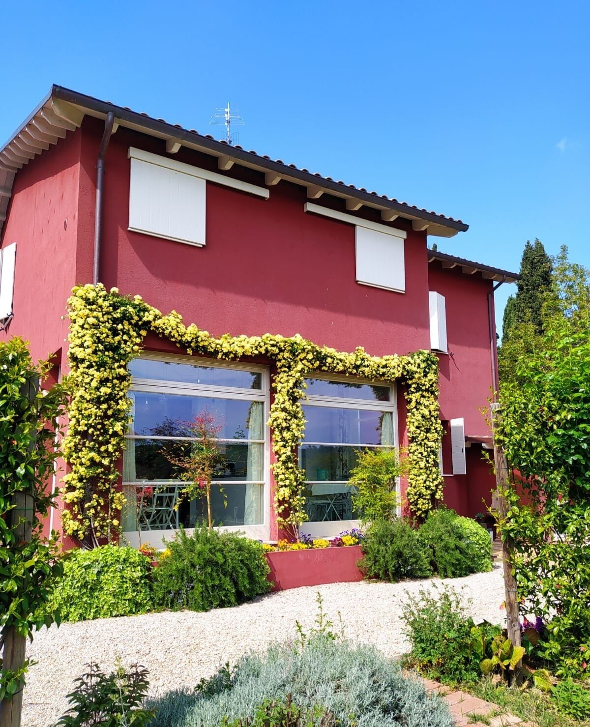 Un edificio rojo con amplias ventanas, una puerta blanca y un jardín con diversas plantas y flores.