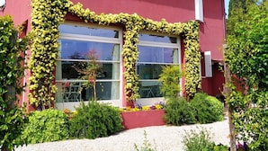 Ein rotes Gebäude mit großen Fenstern, einer weißen Tür und einem Garten mit verschiedenen Pflanzen und Blumen.