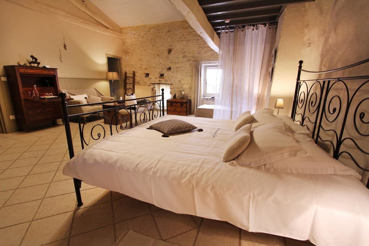 A bedroom with a large bed, a wooden dresser, a nightstand, and a stone wall.
