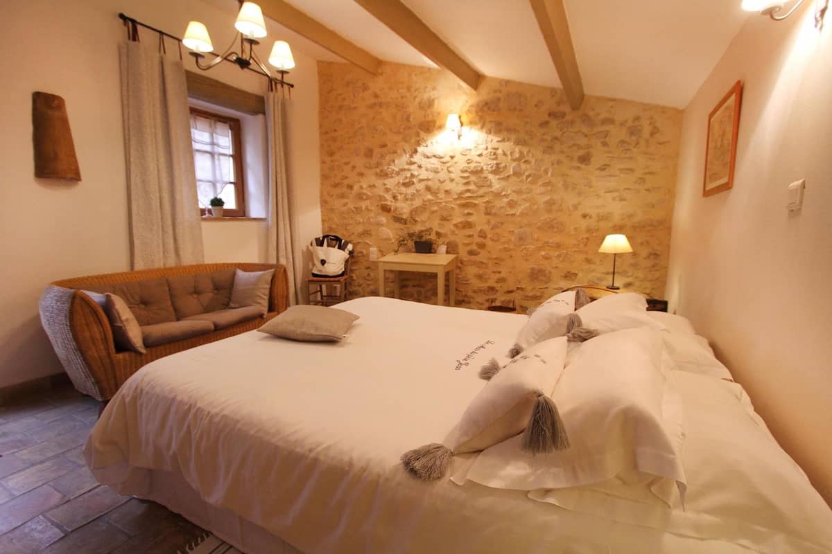 A bedroom with a large bed, a wooden chair, a small table, and a stone wall.