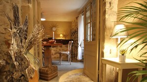 A narrow stone corridor with a rustic interior, a wooden table with a candle, a chair, and a lamp on a small table.