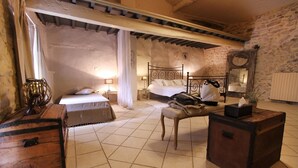 A rustic bedroom with a stone wall, a wooden bed, a chest of drawers, and a bedside table with a lamp.