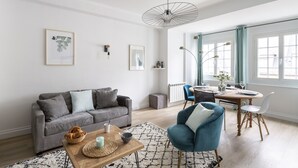 Un saló modern amb un sofà gris, un butaquesa blau, una taula de cafè de fusta i una zona de menjador amb una taula rodona i cadires.