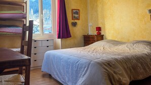 Une chambre à coucher comprenant un lit, un bureau avec une chaise, une fenêtre avec des rideaux, une table de chevet avec une lampe et un tableau accroché au mur.