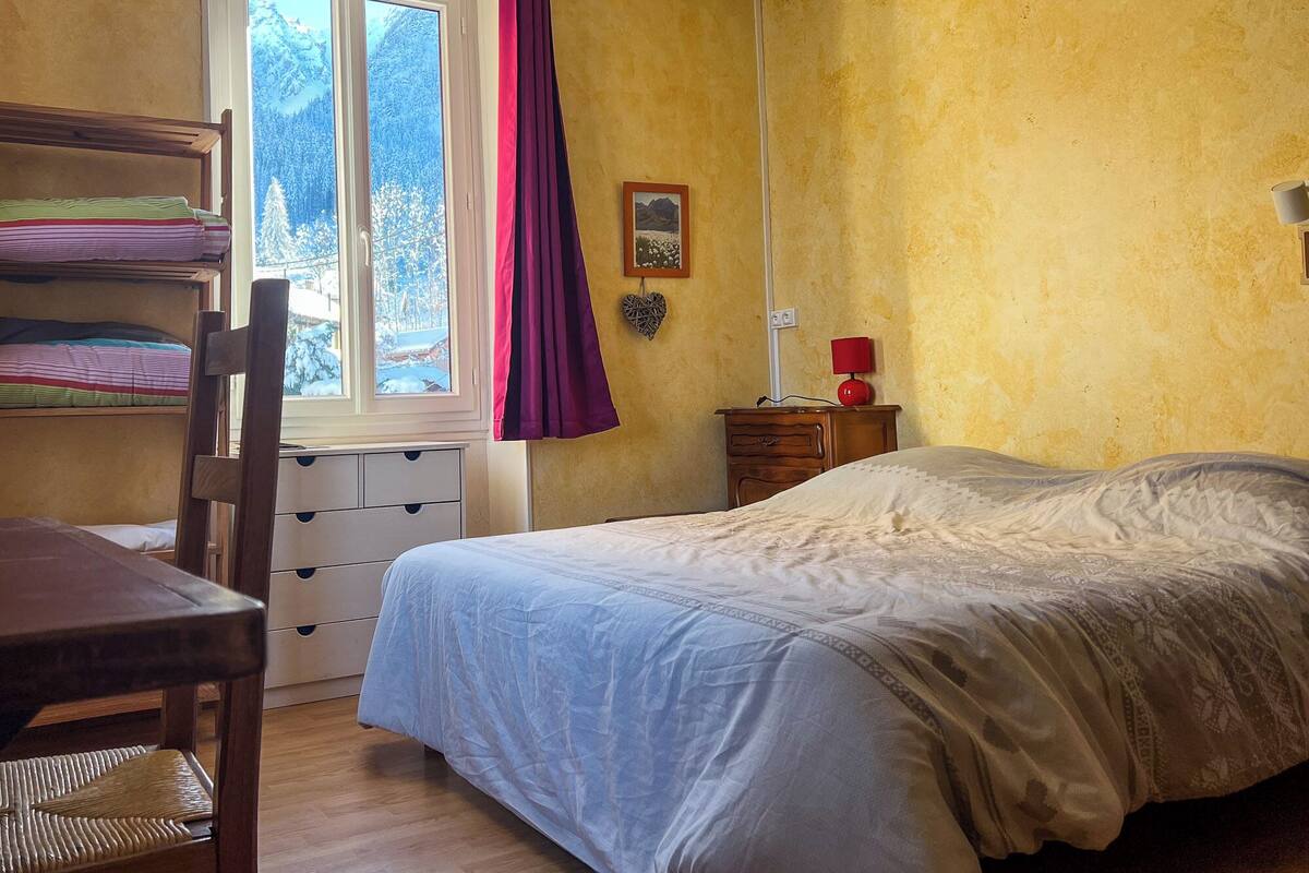Un dormitori amb un llit, una taula de treball amb una cadira, una finestra amb cortines, una taula de nit amb una làmpada i un quadre penjat a la paret.