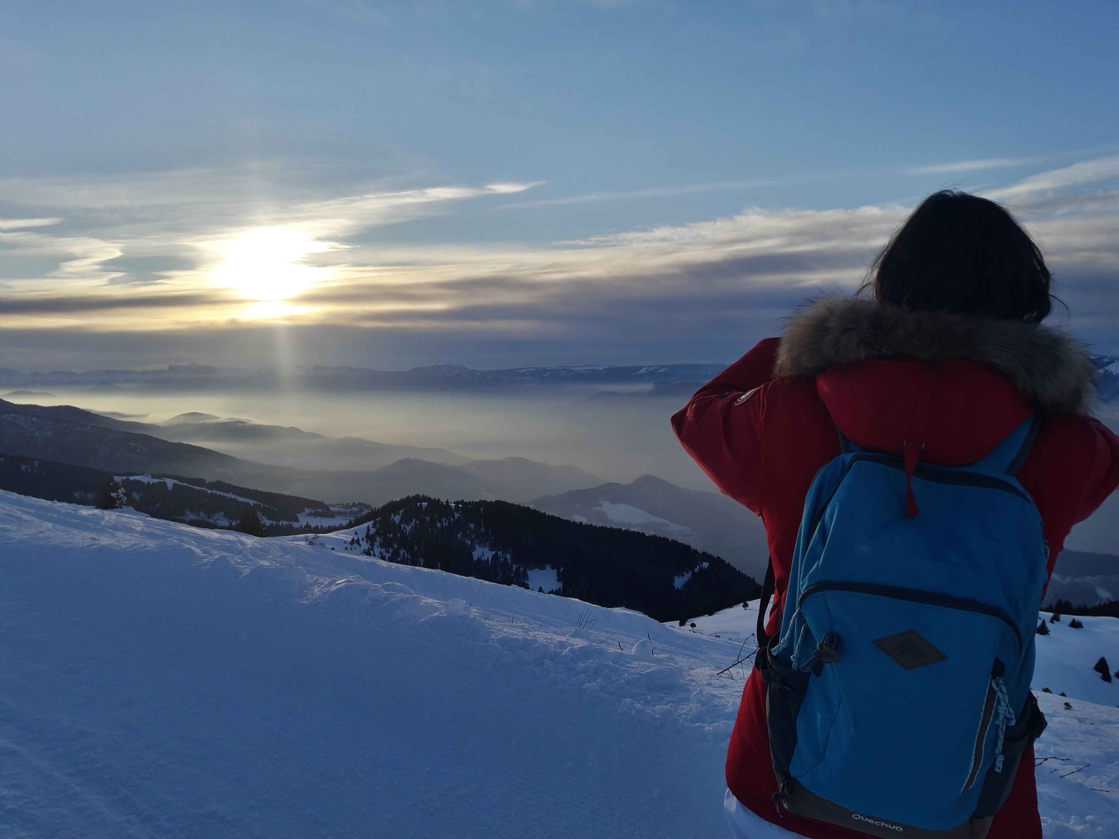 Una persona con una mochila azul en una ladera nevada de montaña, contemplando una cordillera y un amanecer.