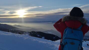 Una persona con una mochila azul en una ladera nevada de montaña, contemplando una cordillera y un amanecer.