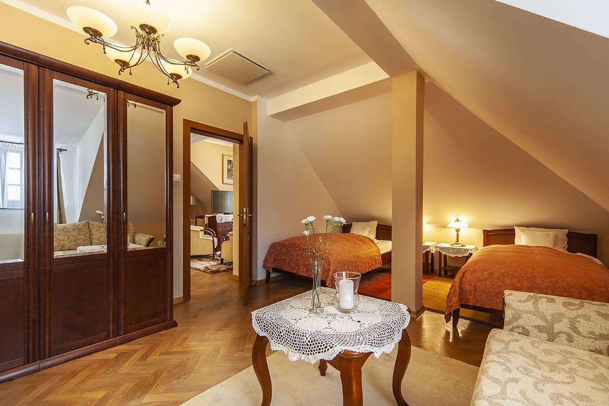 A bedroom with two beds, a wooden wardrobe, a table with a vase and candle, and a chandelier.