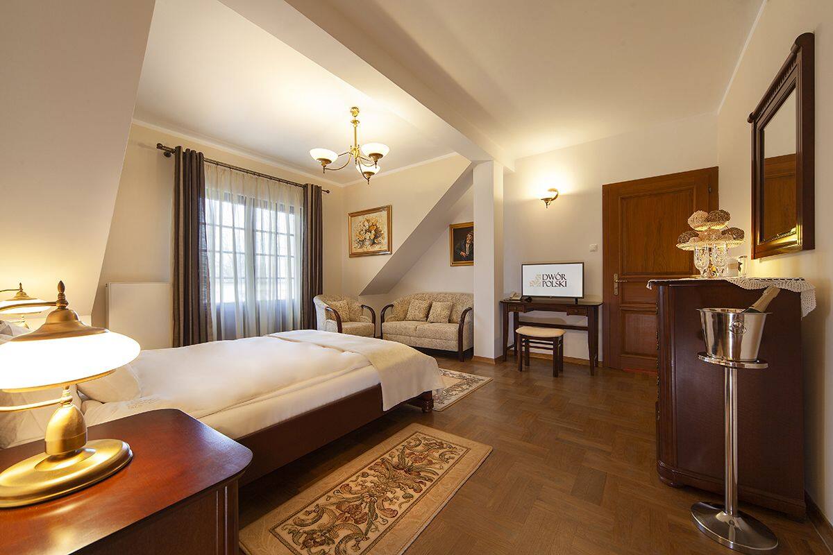 A hotel room with a bed, a desk, a chair, a lamp, a mirror, and a fireplace.