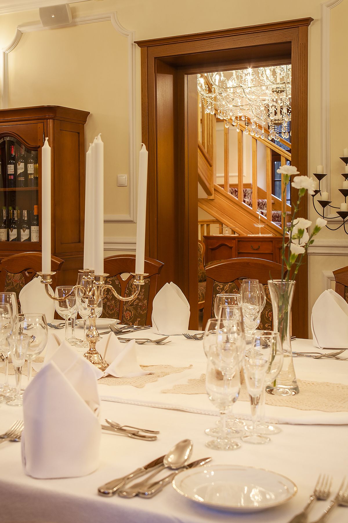 A formal dining room with a long table set for a meal, featuring white tablecloths, glassware, and candles.