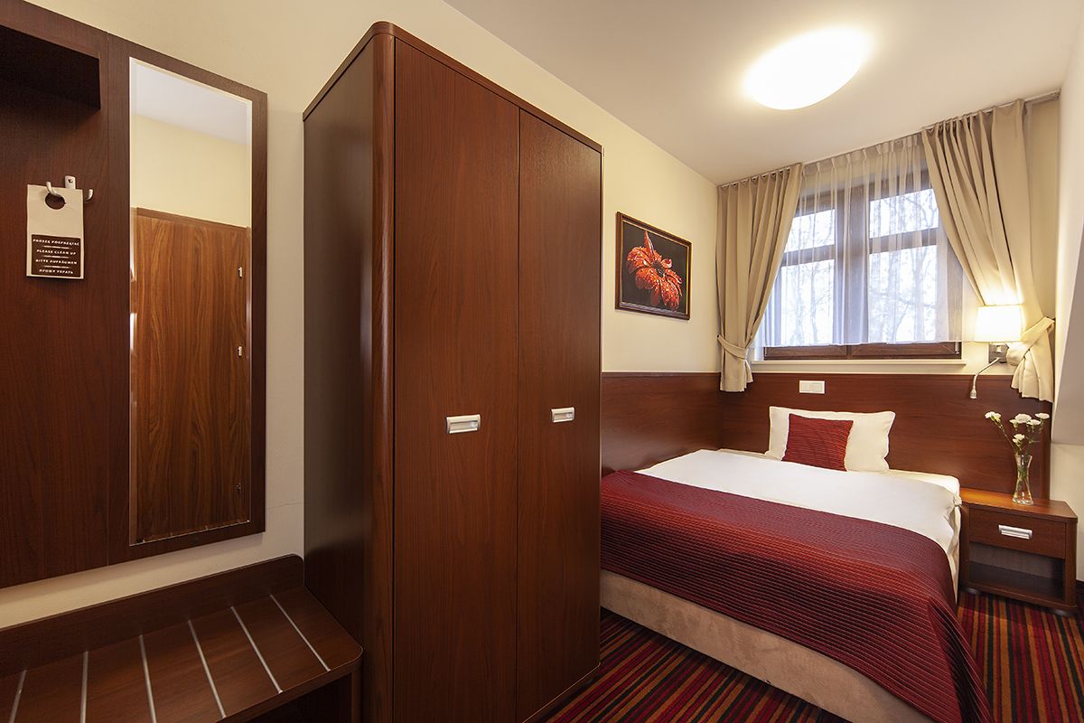 A hotel room with a large wardrobe, a bed with a red blanket, a nightstand with a lamp, and a window with curtains.