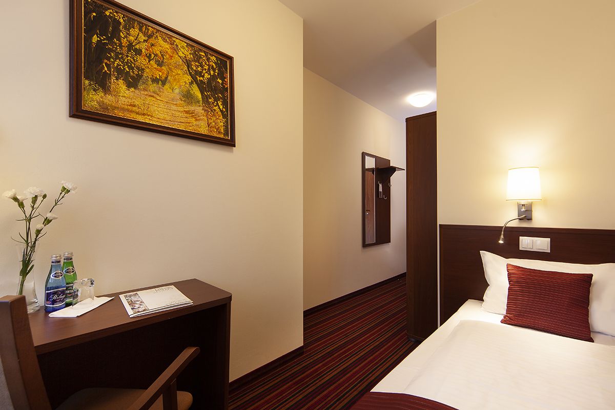A hotel room with a bed, a desk with a chair, a painting on the wall, and a lamp on the bedside table.