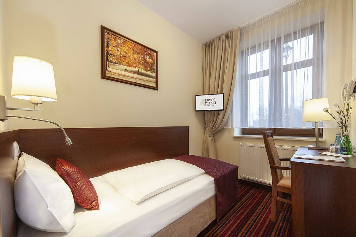 A hotel room with a bed, a desk, a chair, a lamp, a picture on the wall, and a window with curtains.