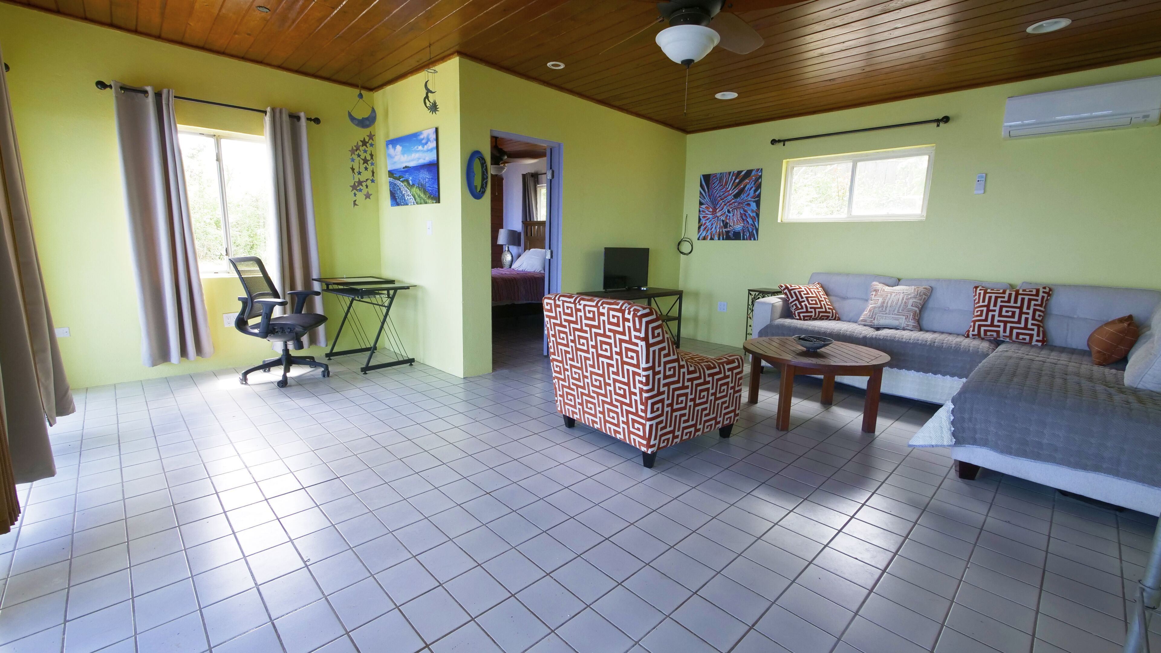 A room with a wooden ceiling, a sofa with patterned cushions, a desk with a chair, and a bed in the background.