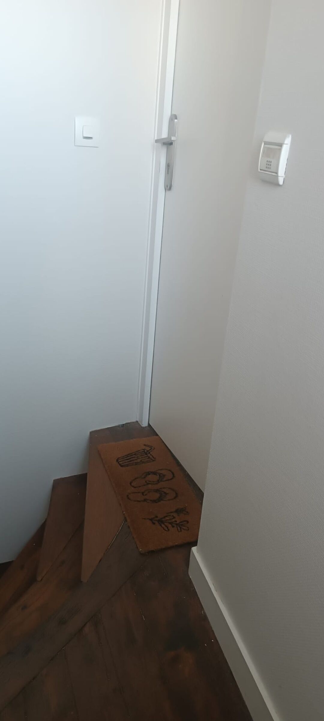Un couloir étroit doté d’un plancher en bois, d’une porte blanche et d’une petite boîte en carton ornée de symboles.