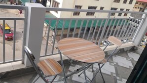 Un balcón con dos sillas y una mesita, con vistas a una calle con coches aparcados y edificios al fondo.