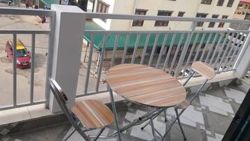 A balcony with two chairs and a small table, overlooking a street with parked cars and buildings in the background.