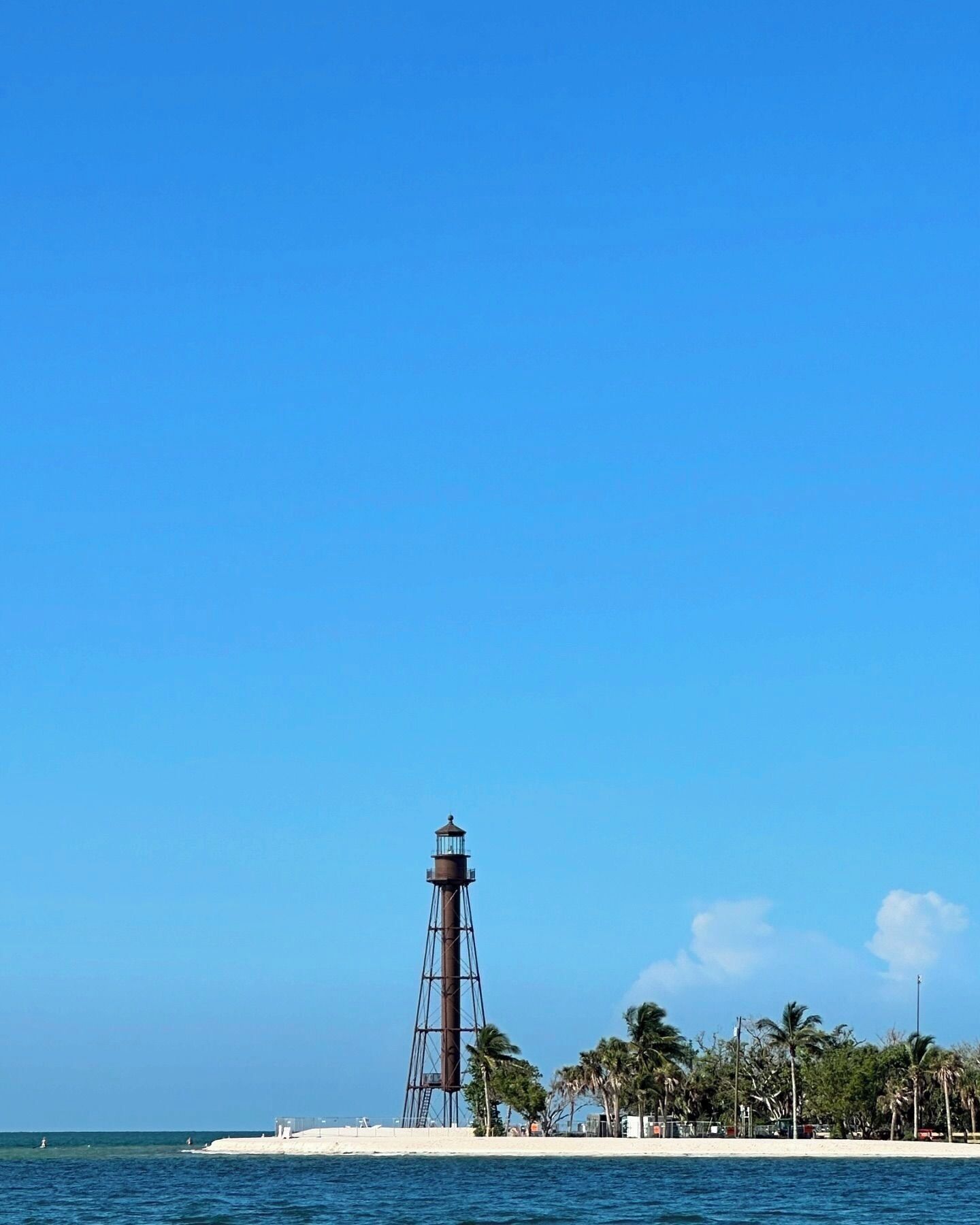 Un faro en una playa arenosa con palmeras y un cielo azul despejado.