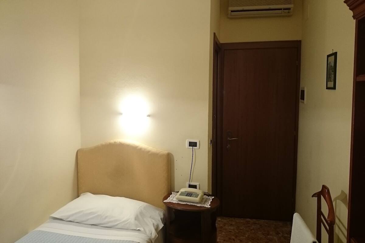 A small, single-bed room with a headboard, a bedside table with a lamp, and a door.