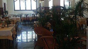 A spacious dining area with high ceilings, large windows, and a chandelier. Tables are set with white tablecloths, and there are potted plants and wicker chairs. - Hotel Pescetto (Albenga)