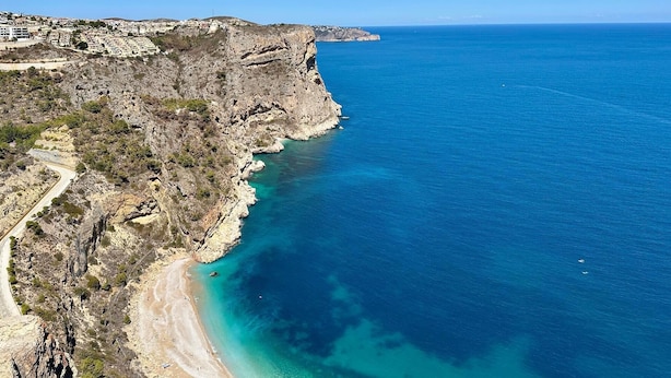 A coastal landscape with a rocky shoreline, a clear blue sea, and a road curving along the coast.