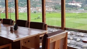 A dining area with a large wooden table and chairs, large windows offering a view of a green landscape with buildings. - Spirit of Bhutan Resort (Paro)