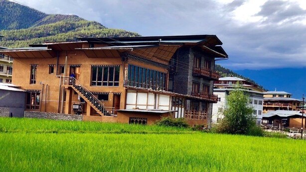 A traditional building with a staircase leading to a balcony, surrounded by lush green fields and mountains in the background. - Spirit of Bhutan Resort (Paro)