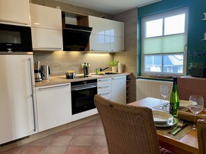 Una cocina moderna con armarios blancos, horno empotrado y comedor con sillas de mimbre y mesa para dos.